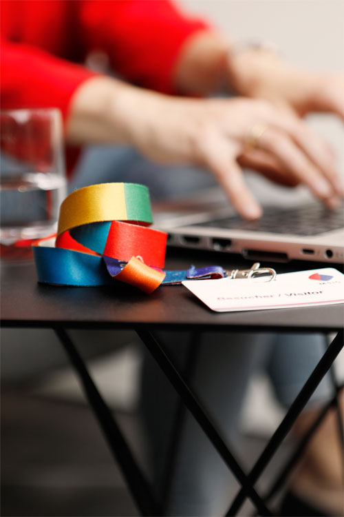 Im Vordergrund Tisch mit Glas, Schlüsselband in Unternehmensfarben und einer Zugangskarte. Im Hintergrund Hände die an einem Laptop tippen.